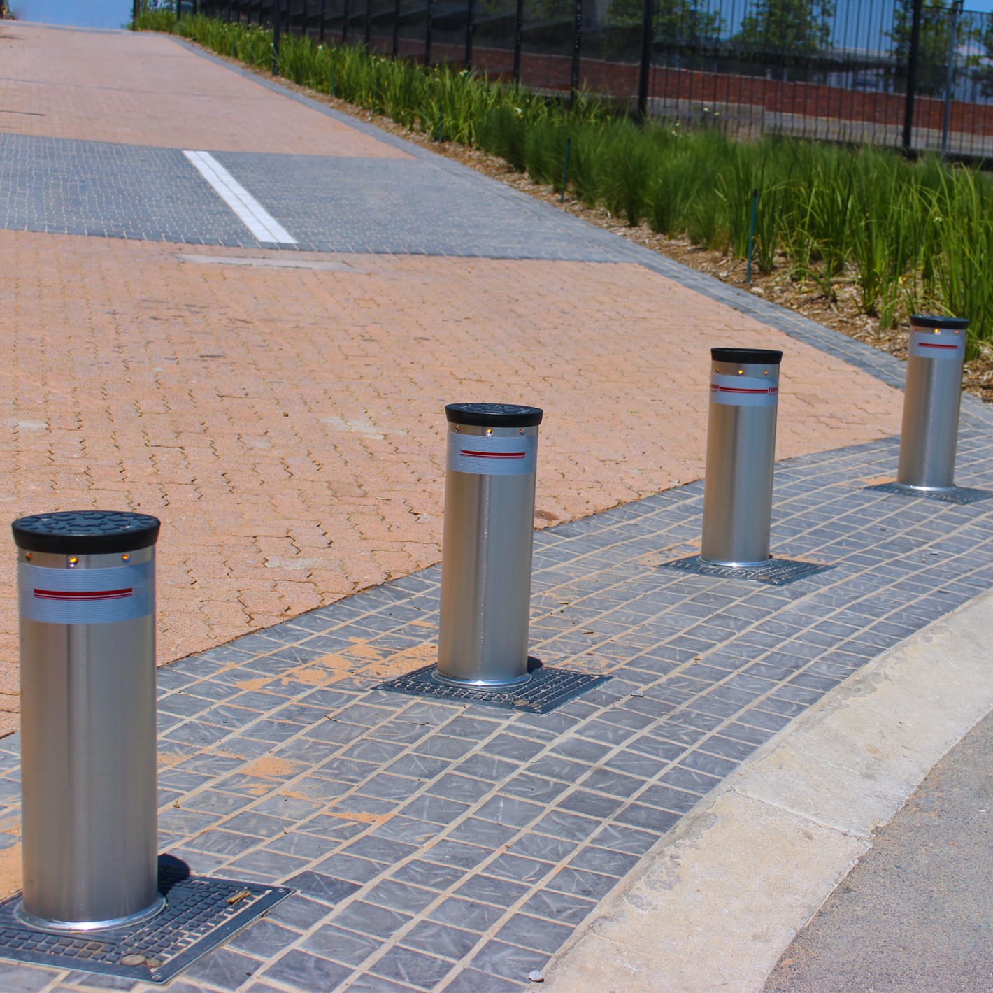 Row of four raised automatic bollards along the edge of a paved driveway.