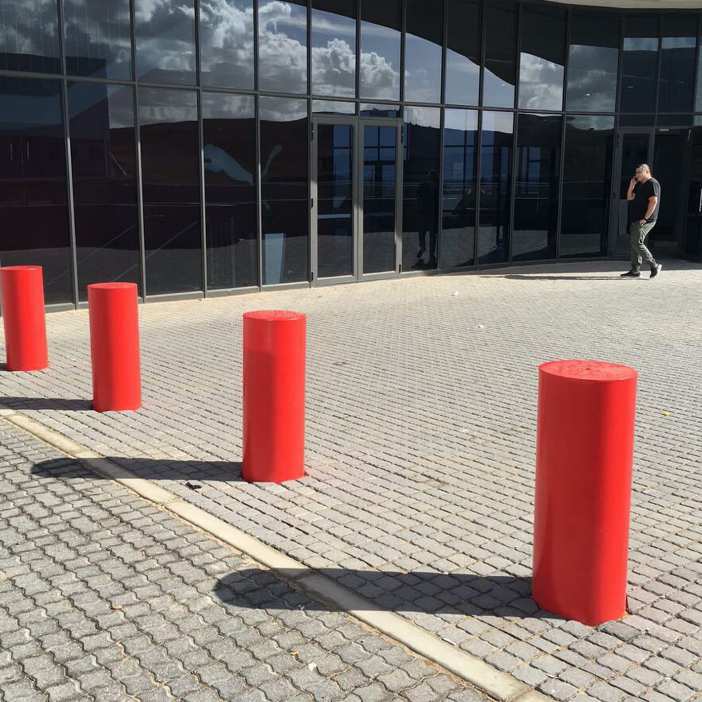 Large red fixed bollards set in block paving in front of a glazed building entrance.