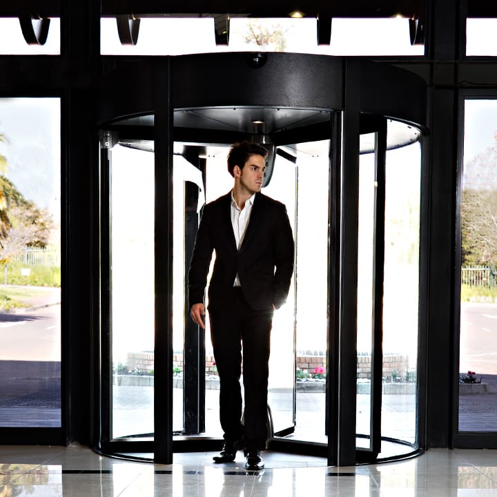 Man in a suit stepping through a black-framed glass revolving door.