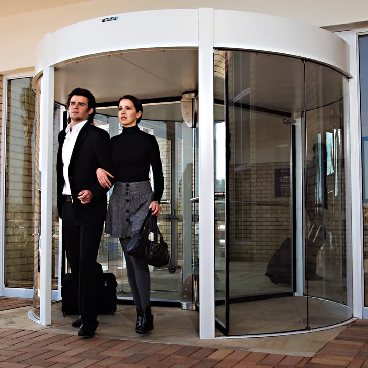 Couple with a travel bag walking out of a white-framed revolving door.