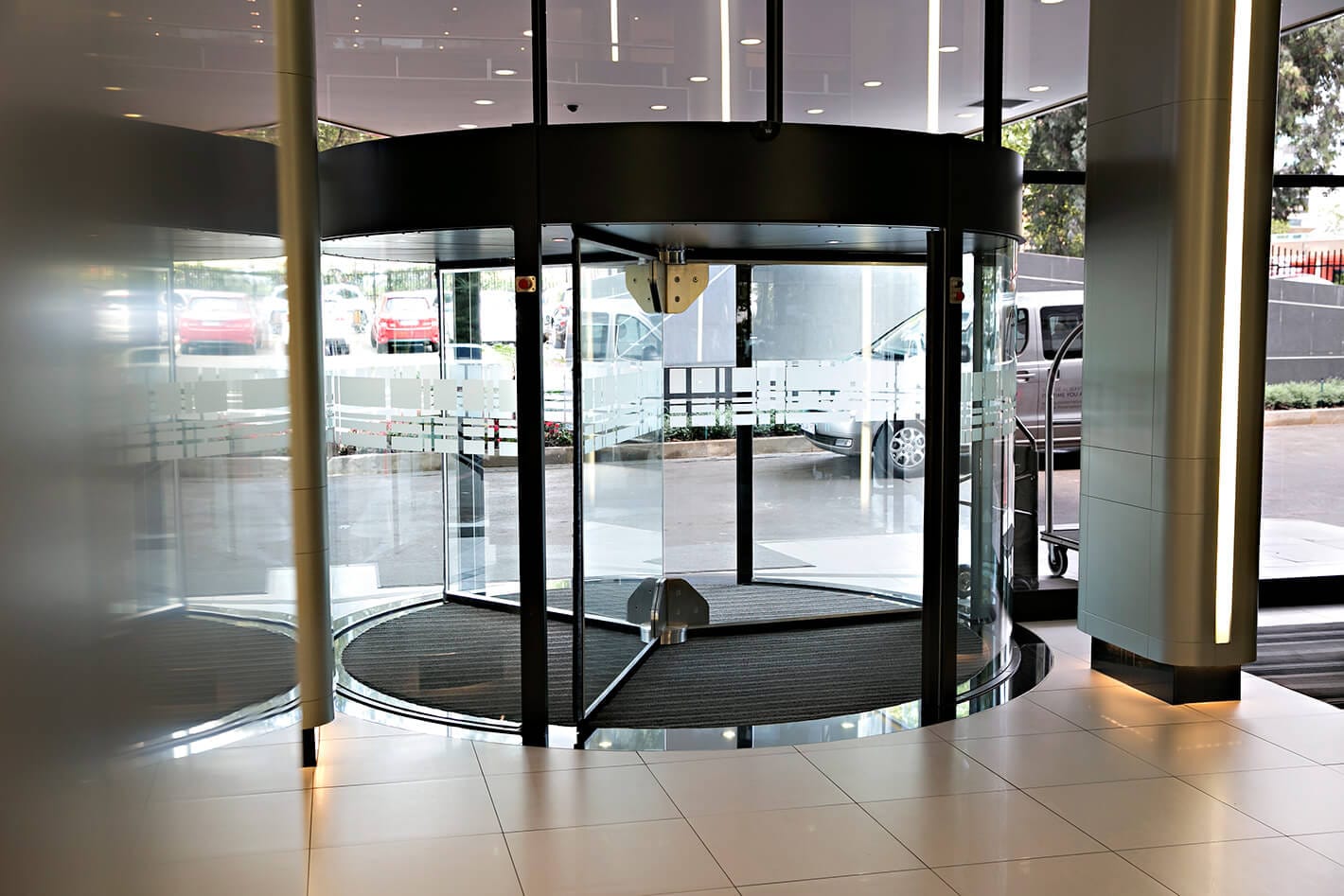 Revolving door with a black canopy in a bright lobby, with daylight and parked cars beyond the glass.