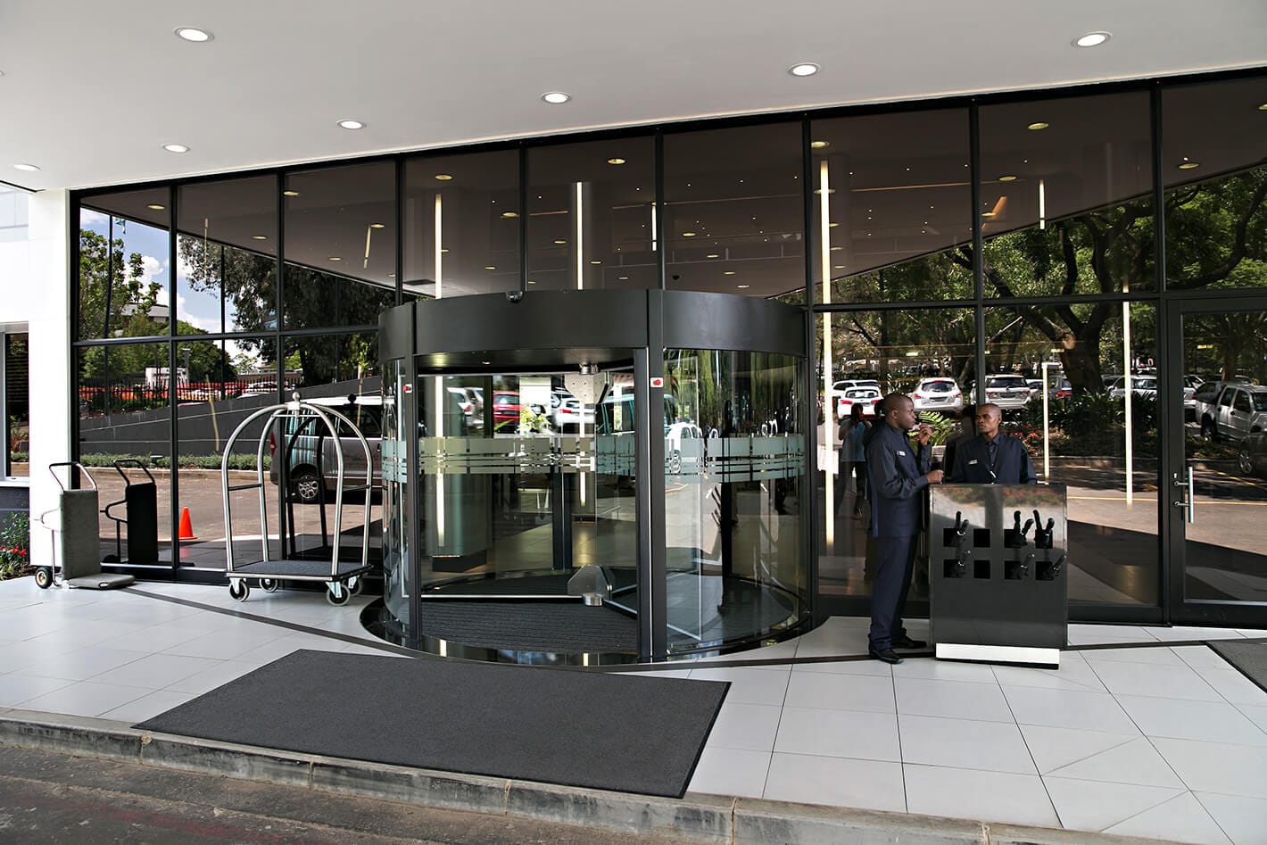 Dark-framed revolving door set into a glass building front, with a lobby and luggage trolley around it.