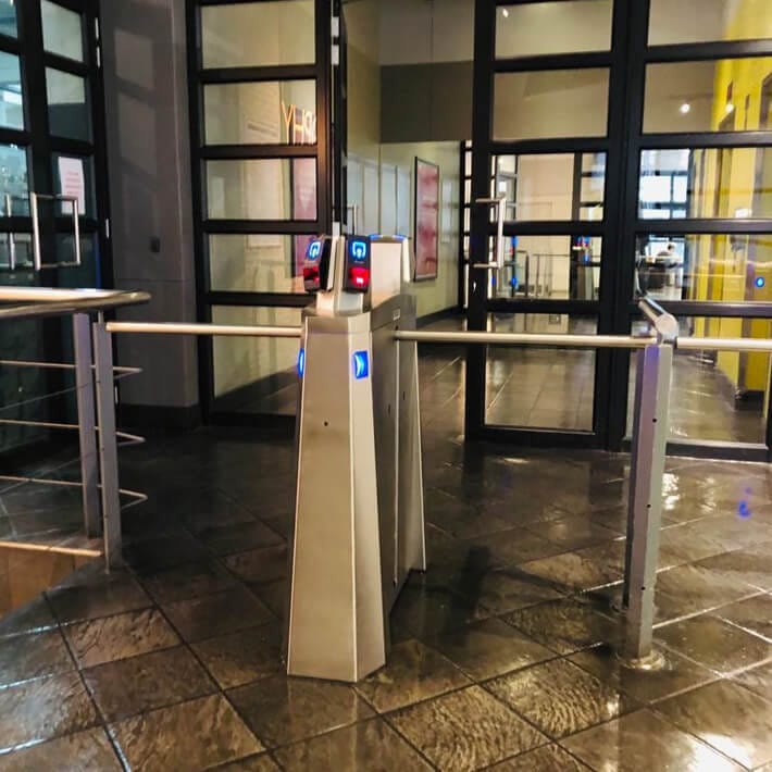 Double Dynamic drop arm barrier installed between glass doors and railings in a building entrance.