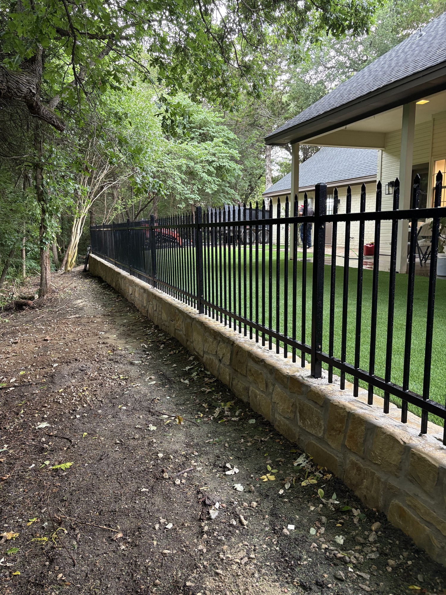 Black pressed point ornamental fence mounted on a stone retaining wall beside a backyard, installed view